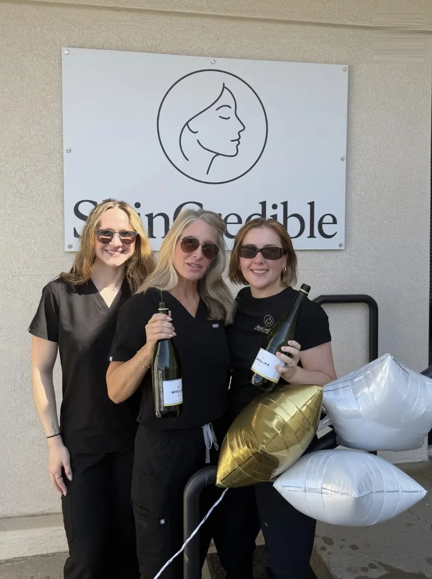 Three members of the Skincredible team holding champagne bottles and gold and white balloons beneath the Skincredible sign outside their North Rock Road clinic in Wichita